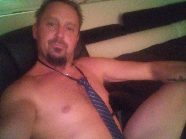 Major-hot, shirtless with a blue striped tie, sitting on a black couch, arms resting, slight smile, short brown hair, goatee, tattoo on right arm, chest visible.