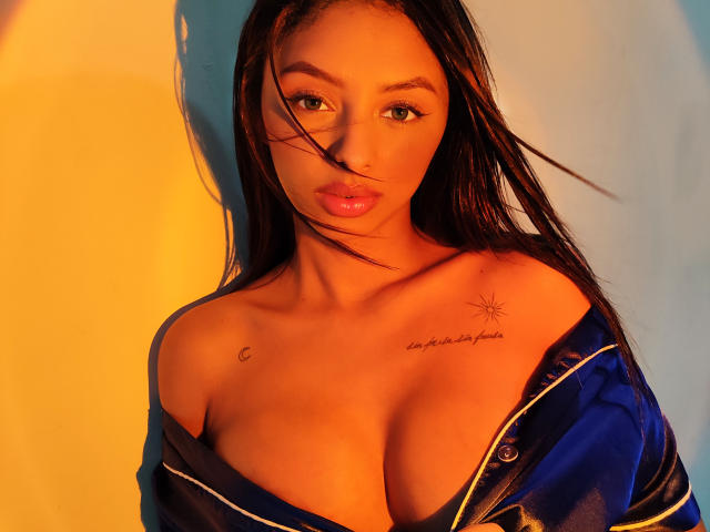 CarlaSofiaDiaz with dark hair, brown eyes, and tan skin, wearing an unbuttoned blue shirt, exposing her large, rounded tits and a small tattoo on her right chest.