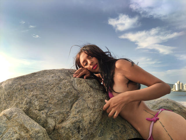 CarlaSofiaDiaz with long black hair, pink bikini, leaning on a rock, showing her small tits and tattoo on her right side, under a blue sky.