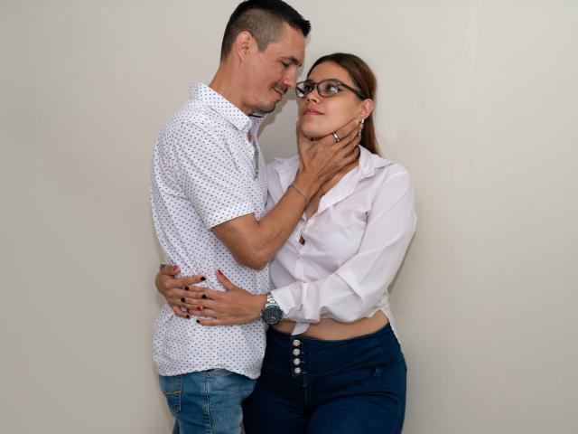 GimenaAndLiam standing, Gimena with glasses, white blouse, blue pants, Liam with short hair, white shirt, blue jeans, Liam's hand on Gimena's neck, Gimena's hand on Liam's waist.
