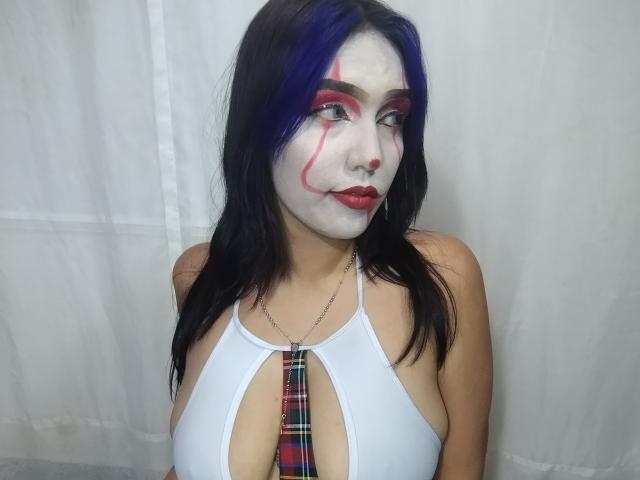 SophieWolf con el rostro pintado de blanco, maquillaje de ojos rojo, cabello negro con reflejos morados, vistiendo un top blanco con cuello halter y una corbata de cuadros, y un piercing en la nariz, frente a una cortina blanca..