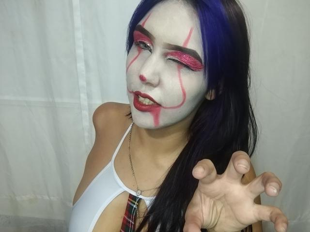 SophieWolf com maquiagem branca no rosto, maquiagem vermelha nos olhos e cabelo azul, piscando com a língua de fora, esticando-se para frente, vestindo uma blusa branca com uma gravata xadrez..