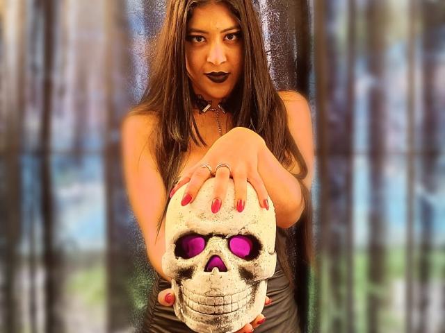 MariaInLove with long brown hair, dark lipstick, and red nails, holding a skull with purple eyes, wearing a black choker and dress, reaching forward with her hands.