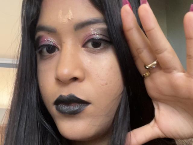 MariaInLove with black lipstick, purple glittery eyeshadow, and maroon nails, showing a hand with gold rings, her face has a small blemish and she has long black hair.