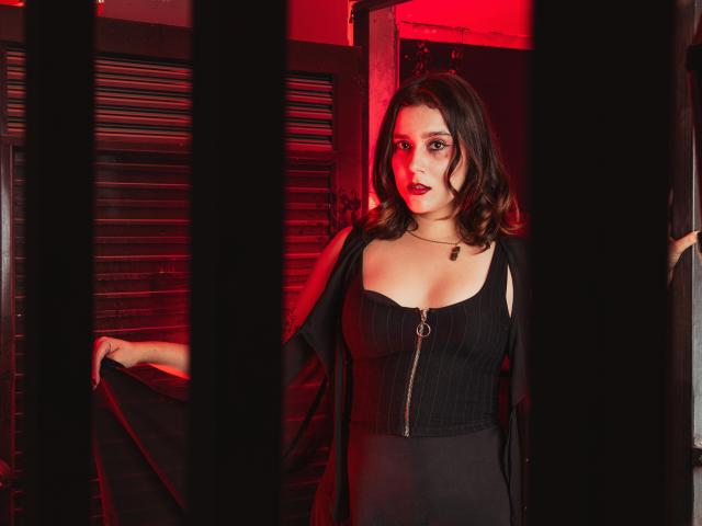 GoddessBar with dark brown hair, red lipstick, and black dress with zipper, holding black cloth, standing between black bars, red light in background, looking forward with confident expression.