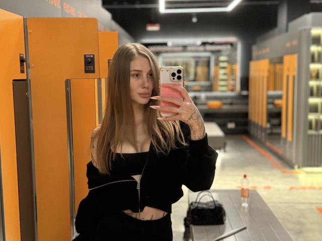 SensualMila mit langen, braunen Haaren, einem schwarzen Top mit einem Schulterauschnitt und schwarzen Hosen, macht ein Selfie vor einem Spiegel in einer Umkleidekabine und zeigt dabei Tattoos an ihrer Hand. Sie hält ein rosa Handy..