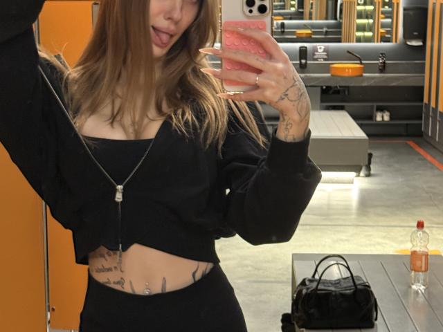 SensualMila with medium tits, tattoos on her stomach and hand, black crop top, black pants, tongue out, holding pink phone, necklace, black jacket, bag on counter, orange wall.