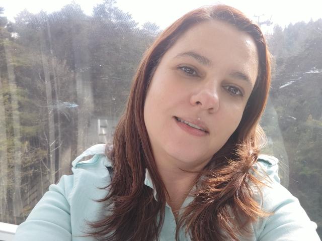AmeliaWilson with medium brown hair, light skin, wearing a light blue hoodie, slight smile, small metal nose ring, standing by a window with forest outside.