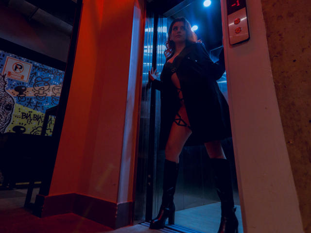 ZoeeMar stands in a doorway, wearing a black coat, strappy lingerie, and thigh-high boots, with her hands on the glass, her tits barely covered, under blue and red lights.