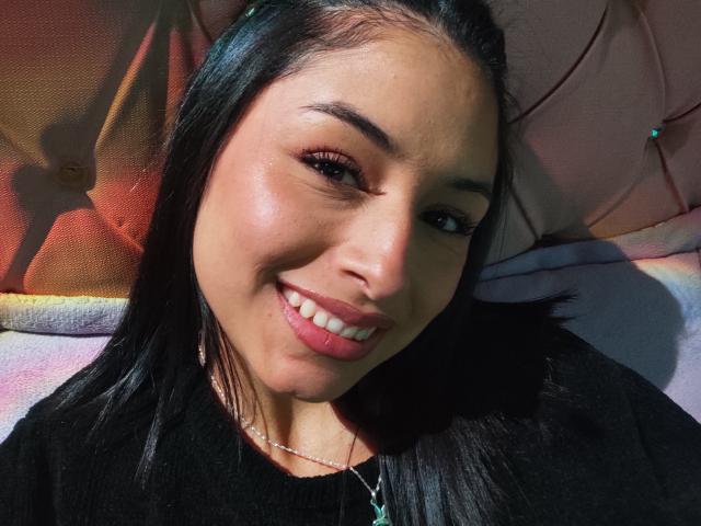 EmilyVv with shiny brown eyes, black hair, and pink lipstick, smiling at the camera, wearing a black top and a thin necklace, lying on a bed.