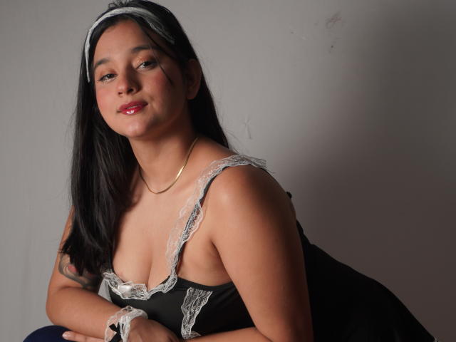 MiaEverly with black hair, wearing a lace-trimmed black dress, gold necklace, and white headband, smiling slightly, showing small tits, and a cute, sexy look.