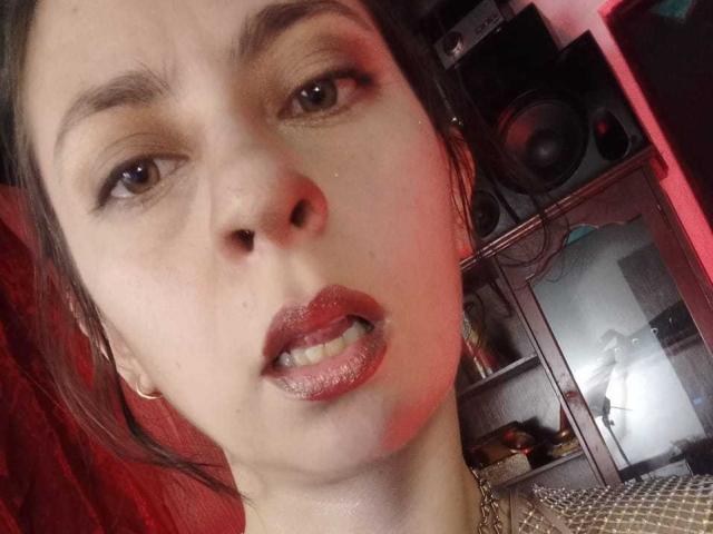 SerenaXYZ heeft glanzende, rode lippen, die lichtjes open zijn en tanden laten zien, met donkerbruine ogen en een lichte frons. Ze draagt gouden oorbellen en heeft een rozeachtige gloed op haar gezicht..