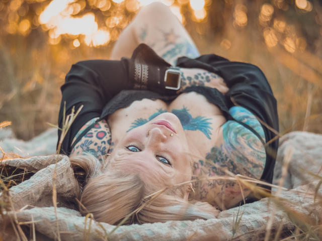 Tattooed MsGoodGood with blonde hair, blue eyes, and nose ring, lying on grass, wearing black top, belt, and lace, looking up, with visible boobs and tattoos.