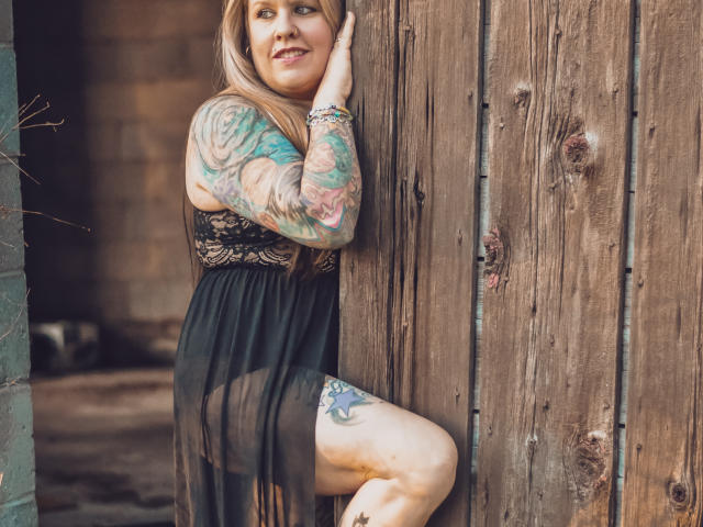 MsGoodGood, com tatuagens no braço e na coxa, vestindo um vestido preto transparente, sorrindo, encostada em uma parede de madeira, com a perna levantada, mostrando sua vulva..