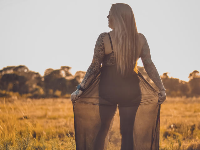 MsGoodGood, com cabelos loiros longos, braços tatuados, vestido preto transparente e nádegas visíveis, está em um campo ensolarado, segurando as laterais do vestido, com o sol se pondo atrás dela..