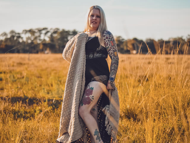 MsGoodGood se tient dans un champ, vêtue d'une robe noire en dentelle, avec des manches à motifs léopard, et enveloppée d'une couverture beige, avec des tatouages sur sa jambe et son bras, souriant à l'appareil photo..