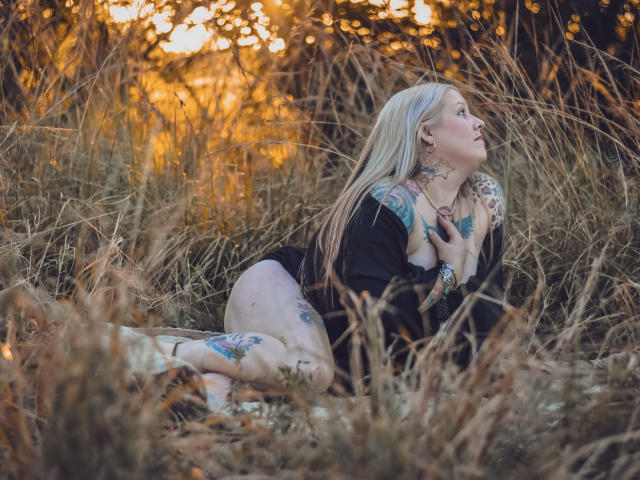 MsGoodGood with blonde hair, tattoos, and black top, kneeling in dry grass, touching her tits, looking up, sun setting in the background, bare ass partially visible, arms and legs tattooed.