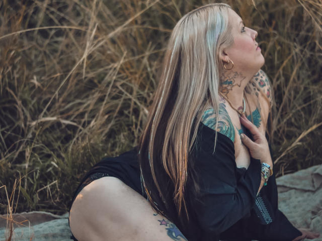 MsGoodGood, com cabelo loiro, tatuagens e uma camisa preta, ajoelhada, mostrando o bumbum, as tatuagens nas costas e olhando para cima. Grama e pedras ao fundo. A mão dela está no peito..