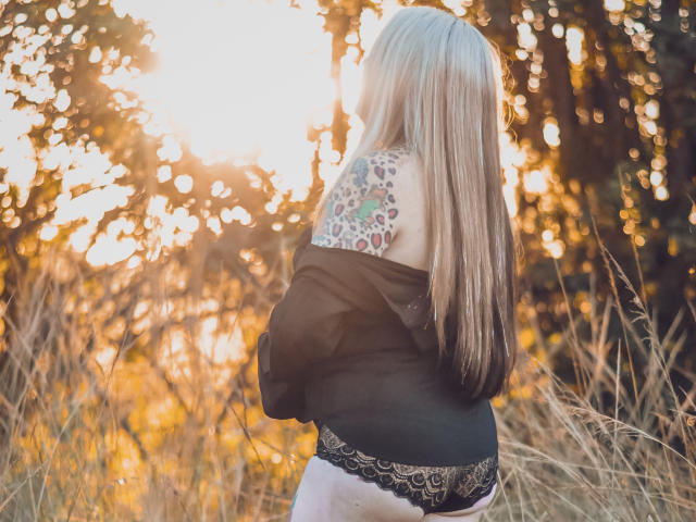 MsGoodGood, con cabello rubio largo, una blusa negra con un hombro descubierto y ropa interior de encaje negra, mostrando un colorido tatuaje de leopardo en la parte superior de la espalda, de pie en hierba iluminada por el sol..