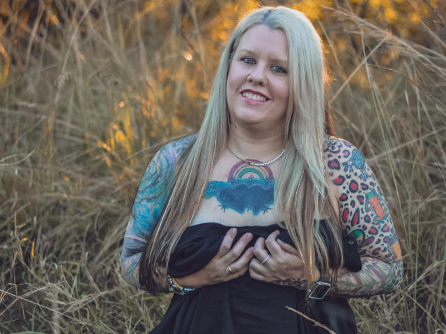 MsGoodGood ha lunghi capelli biondi, tatuaggi sul petto e sulle braccia, ed è sorridente mentre tiene il suo vestito nero, mostrando il seno. Si trova in un campo con erba secca..