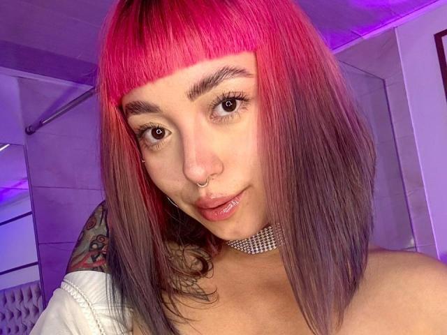 GigiVolkova with pink and purple dyed hair, nose ring, choker, tattoo on shoulder, and lipstick, looking at camera with purple and pink lighting in the background.