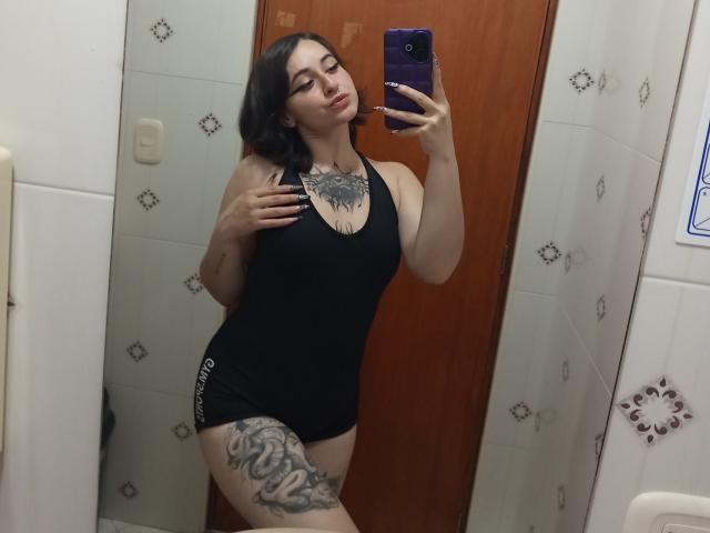 MissTat with black tank top, showing off her tits and a big tattoo on her thigh, taking a mirror selfie with a pouty face, holding a purple phone.