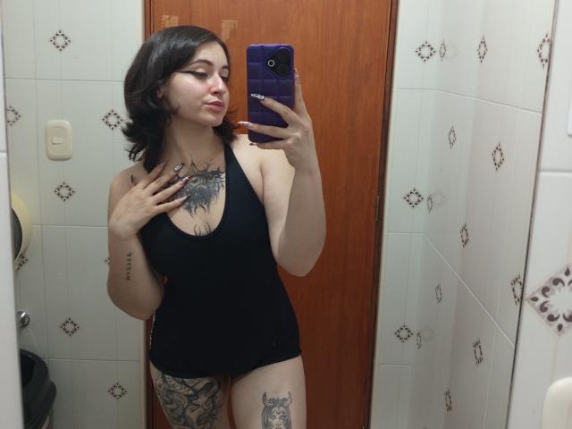MissTat with dark hair, black tank top, tattoos on chest and thigh, holding purple phone, mirror selfie, showing her tits, pussy, and tattoos in a bathroom.