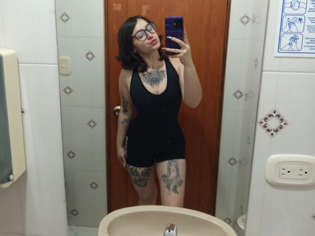 MissTat with glasses, black tank top, and shorts, showing off tattoos on chest, thigh, and arm, taking a mirror selfie with a purple phone, in a bathroom.
