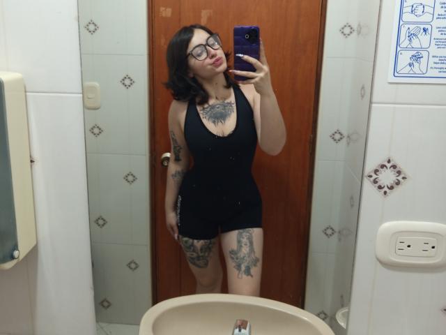 MissTat with glasses, black tank top, and shorts, showing tattoos on chest, thigh, and arm, taking a mirror selfie in a bathroom, holding a purple phone.