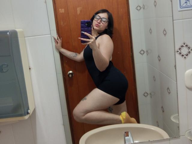MissTat has pale skin, black bob, glasses, black tight dress, yellow socks, tattoos, and is posing in a bathroom, holding a purple phone, with her ass and tits visible.