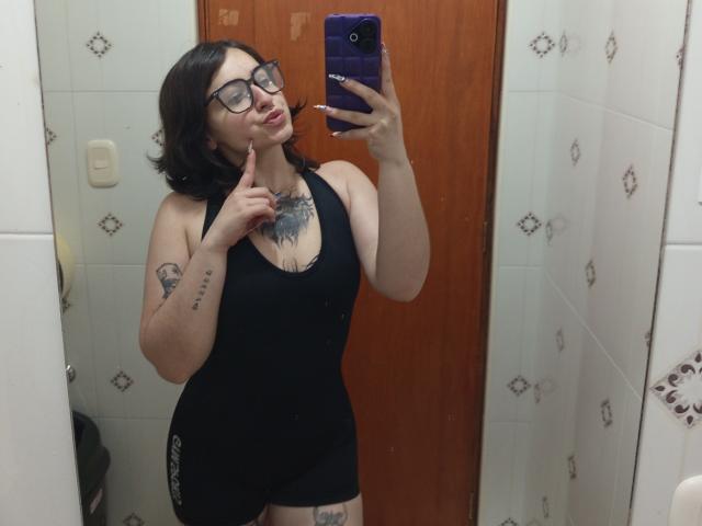 MissTat with black hair, glasses, and tattoos, wearing a black tank top and shorts, holds a purple phone, pointing to her lips, showing her tits and ass in a mirror.