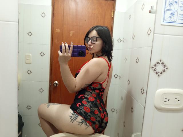MissTat with glasses, black hair, red lipstick, wearing a red and black kiss-patterned dress, showing her ass and a tattoo on her thigh, taking a mirror selfie.