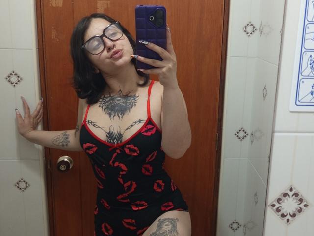 MissTat with black hair, glasses, and tattoos, wearing a red and black kiss-print dress, taking a mirror selfie in a bathroom, showing her tits and thigh tattoo.