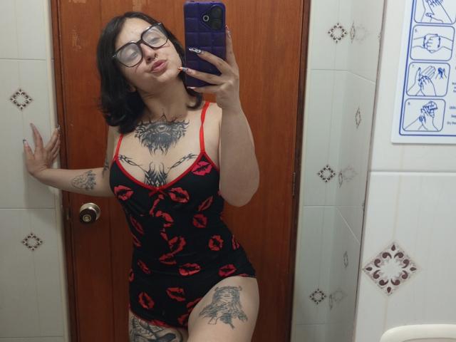 MissTat with black hair, glasses, red lipstick, black and red kiss-patterned camisole, showing her large tits and tattoos, taking a mirror selfie with a purple phone.