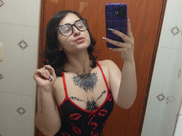 MissTat with glasses, black hair, and red-black dress, showing off chest tattoos, holding purple phone, in mirror selfie. Boobs visible, tattooed chest, fingers with white nail polish.