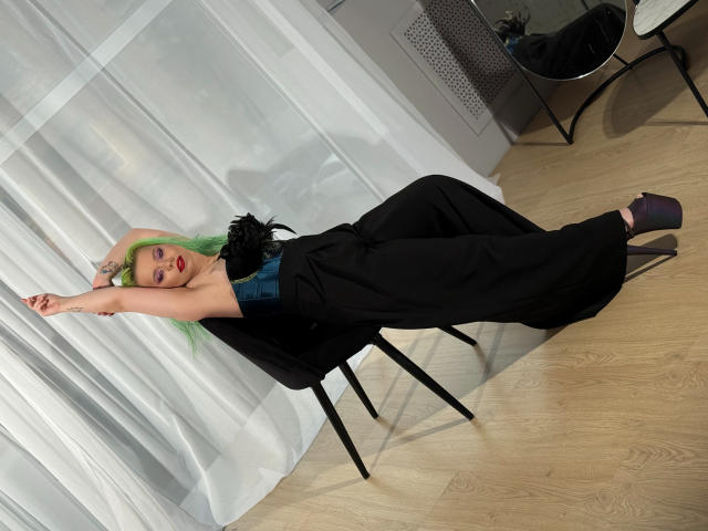 EnergyBurnny with green hair, arms stretched, wearing black pants, blue top, black heels, and black flower accessory, standing next to black chair on wooden floor.
