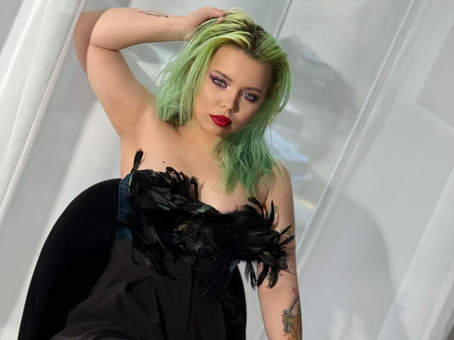 EnergyBurnny with green hair, red lipstick, and black feather dress, showing tits, arm tattoo, and hand in hair, looking at camera.