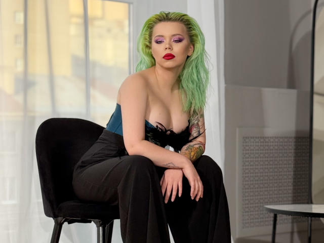 EnergyBurnny with green hair, red lipstick, purple eyeshadow, black strapless top showing tits, black pants, sitting on black chair, tattooed arm, confident expression.