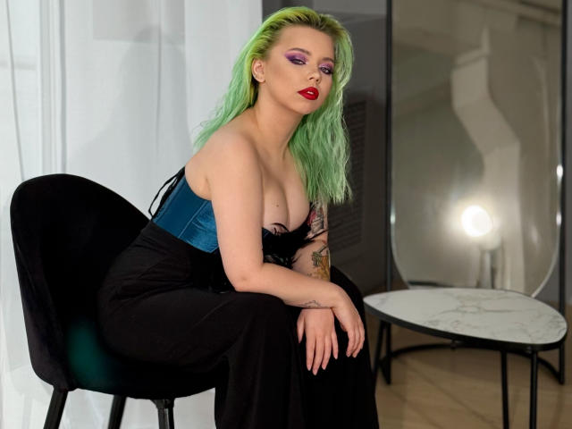 EnergyBurnny with green hair, red lipstick, purple eyeshadow, blue velvet top, black lace, and black skirt, sitting on black chair, showing big tits, tattoo on right arm.