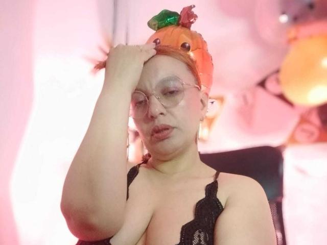 RoxieJackson wearing a pumpkin hat with green and red eyes, glasses, and black lace top, touching her head with a slightly confused expression. Her tits are visible.