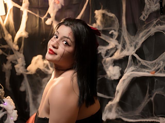 LeylaMiller, with black hair and red lipstick, looks over her shoulder, showing her bare shoulders and black top, surrounded by white smoke. Her red hair clip stands out.