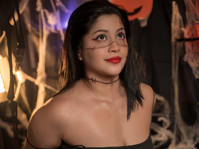 Leyla Miller, met donker haar, rode lippenstift, een zwart top met open schouders en een zwarte choker met stiksels, kijkt omhoog met een lichte glimlach, waarbij haar borsten en schattige gezicht zichtbaar zijn..