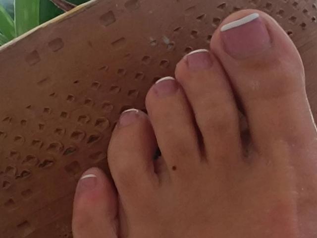 PennySweetLane-hot's foot with pinkish-brown skin, white French tips, and a small mole on the big toe, resting on a brown perforated surface.