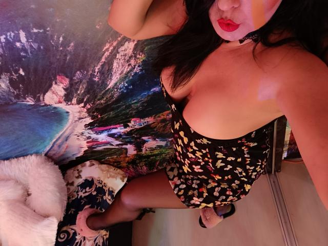 AmberSunshine with medium tits, wearing a black floral dress, choker, and pink heels, has dark hair and red lipstick, standing in a room with a colorful beach painting.