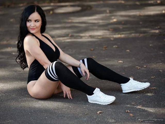 Sydney-Pearl-un costume intero nero, aderente, con scollatura profonda a V, che mette in mostra il seno, calze nere a ginocchio con strisce bianche, scarpe da ginnastica bianche, seduta sull'asfalto, capelli lunghi neri, sorridente, sexy..