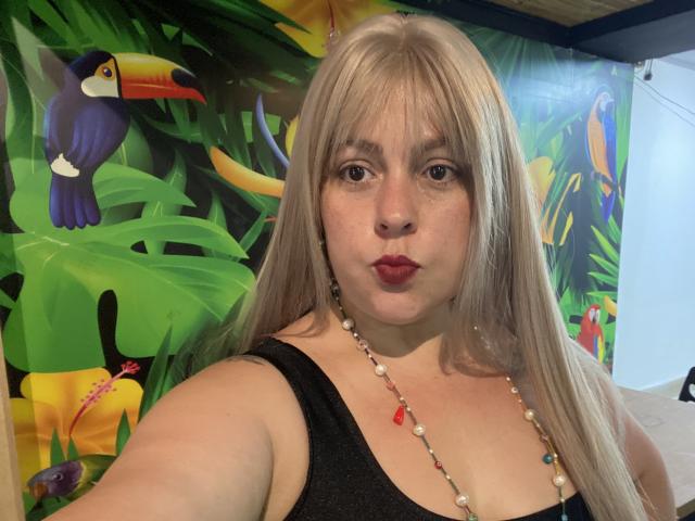 SamanthaGold with long blonde hair, red lipstick, black tank top, and beaded necklace, showing her tits in a selfie. Tropical bird and plant mural in the background.