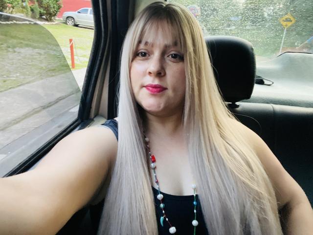 SamanthaGold with long blonde hair, pink lipstick, and a beaded necklace, sitting in a car, taking a selfie, showing her boobs in a black top.
