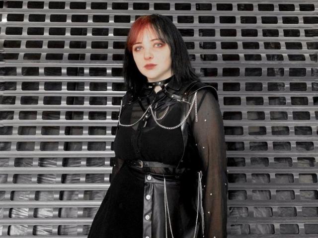 SweetLilu with pale skin, black hair, and red highlights, wearing a sheer black dress with chain details and buttons, standing against a grid metal wall.