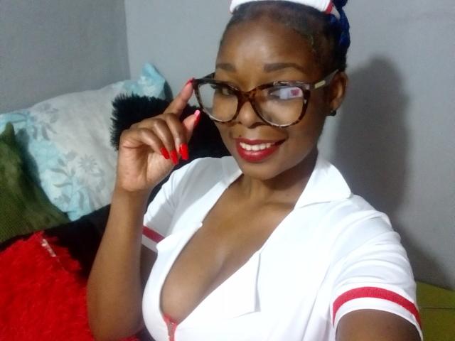 LadyLluck with dark skin, red lipstick, and glasses, wearing a low-cut white nurse outfit, touching her glasses, showing her cleavage, and smiling. Her red nails match her outfit.