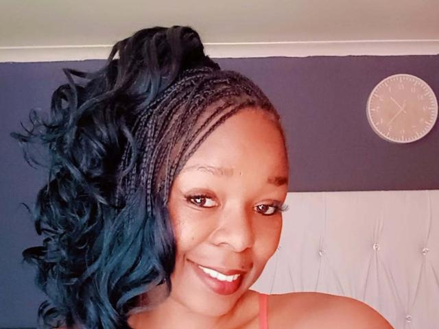 LadyLluck with dark blue curly hair, braided front, smiling, pink lipstick, brown eyes, wearing pink top, clock on dark wall, white headboard, cute and attractive.
